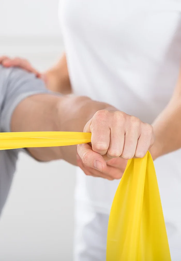 Mann betreibt allgemeine Physiotherapie und macht Armübungen mit einem Theraband. Lucrezia Blatzheim unterstützt die Armübung als Physiotherapeutin.